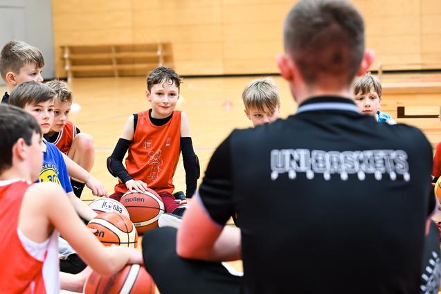 Gruppe von Jungen beim Basketballtraining, sitzend auf dem Boden mit Bällen, Trainer spricht zu ihnen.