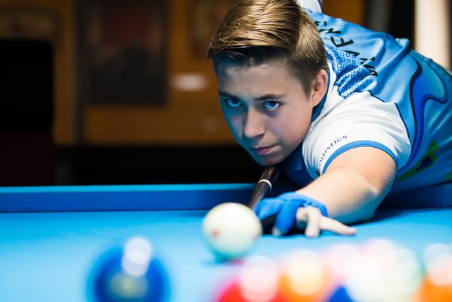 Junge spielt Poolbillard, konzentriert auf die weiße Kugel, umgeben von bunten Bällen auf einem blauen Tisch.