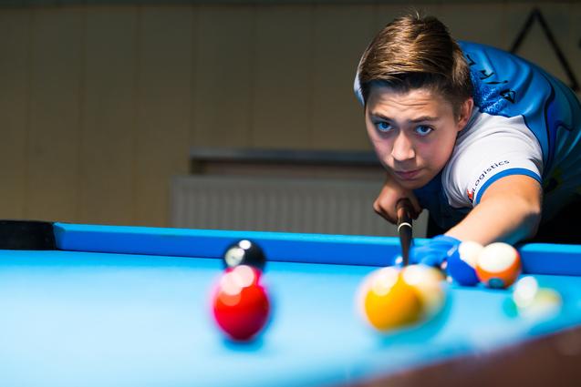 Junger Spieler konzentriert sich auf einen Stoß mit einem Queue an einem Billardtisch, farbige Kugeln sind sichtbar.