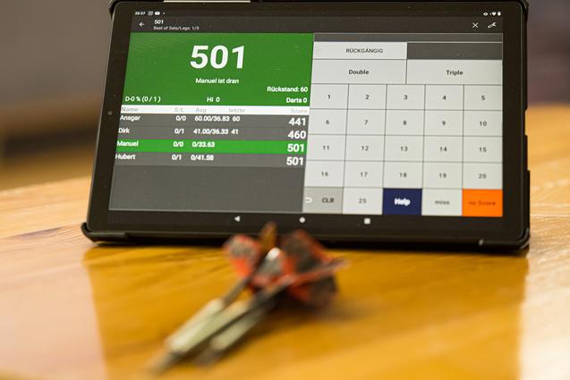 Tablet mit Dartspielstand und Pfeilen auf einem Tisch, im Hintergrund ein Spieler beim Dartwerfen.