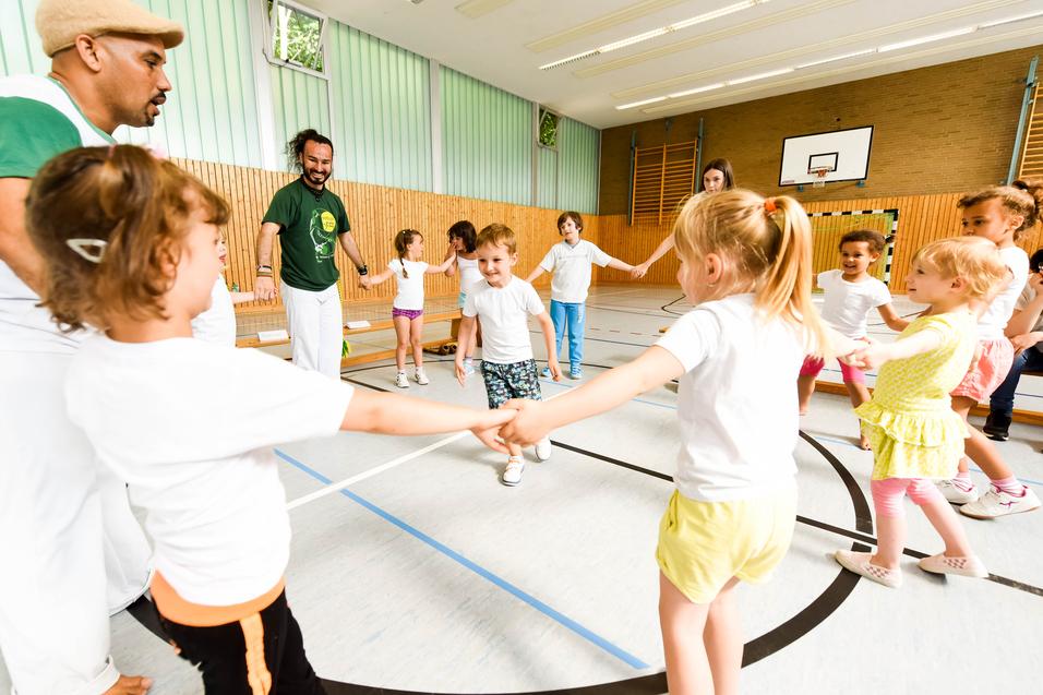 Gruppe von Kindern in T-Shirts und Erwachsenen in einer Turnhalle, die gemeinsam einen Kreis bilden und spielen.