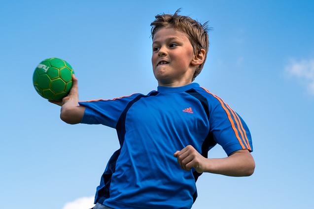 Junge springt auf einem Sportplatz und wirft einen grünen Handball, umgeben von einem Zaun und Bäumen im Hintergrund.