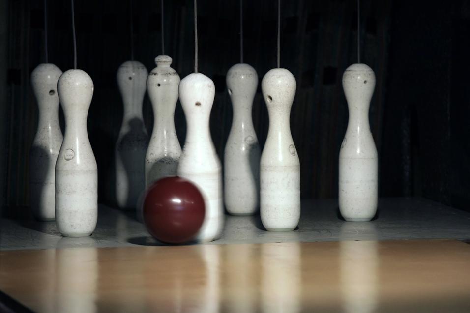 Eine rotbraune Bowlingkugel rollt auf eine Reihe weißer Kegel auf einer Bowlingbahn zu.