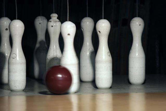 Eine rotbraune Bowlingkugel rollt auf eine Reihe weißer Kegel auf einer Bowlingbahn zu.