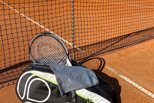 Tennisplatz mit Netz, einer ARTENGO-Tasche und einem Schläger im Vordergrund auf dem rotem Sand.