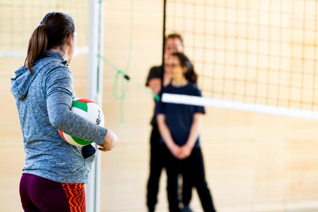 Eine Person mit einem Volleyball steht vor dem Netz, während zwei weitere Personen im Hintergrund warten.