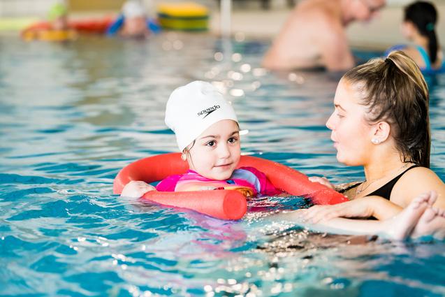 Kind mit Schwimmhilfe im Wasser, unterhält sich mit einer Erwachsenen, beide lächeln. Hintergründe zeigen andere Schwimmer.